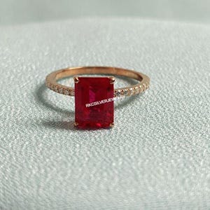 Puede incluir: Un anillo de oro rosa con una gran gema roja rectangular. La banda está incrustada con pequeñas piedras transparentes. El texto "RKCSILVERJEWELRY" es visible en la gema. El anillo está sobre un fondo azul claro.