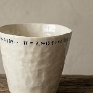 May include: A handmade, off-white ceramic cup with a textured surface. The cup features the mathematical constant Pi, with the digits written in blue ink around the rim. The cup sits on a rustic wooden surface.