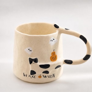 May include: A cream-colored ceramic mug with a speckled finish, featuring a black and white cat illustration, two small white ghosts, and an orange pumpkin. The handle has black spots, and the text HALLOWEEN is printed with a pumpkin for the O.
