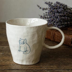 May include: A cream-colored ceramic mug with a textured, uneven surface and a single handle. The front of the mug features a simple, dark blue line drawing of a cat with wide, circular eyes and a small mouth, sitting in a seated position.