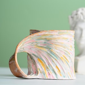 May include: A ceramic mug with a textured, rippled surface featuring a hand-painted pattern of soft pink, pale yellow, and light green brushstrokes. The mug has a wide, curved handle that extends from the base to the rim, finished in a warm brown.