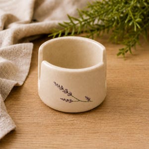 Może przedstawiać: Kremowa ceramiczna miseczka na włóczkę o gładkim, nakrapianym wykończeniu z wycięciem w kształcie litery U z jednej strony. Na zewnętrznej stronie znajduje się delikatna, ciemnofioletowa ilustracja botaniczna gałązki lawendy.