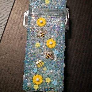 Può includere: Un astuccio per accendino azzurro, decorato con strass iridescenti, perle, fiori gialli e blu e ciondoli a forma di ape. L'astuccio ha una cerniera e una chiusura in metallo argentato.