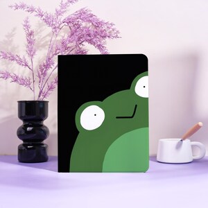 Op de afbeelding: Een zwart notitieboek met een groene kikkerillustratie. De kikker heeft grote witte ogen en staat tegen een zwarte achtergrond. Het notitieboek ligt op een paars oppervlak, met een zwarte vaas en een witte mok met een lepel.