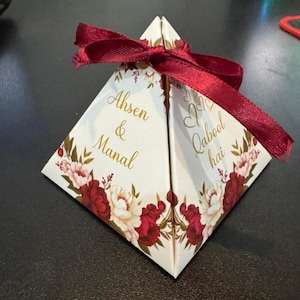 May include: A white, pyramid-shaped gift box with a burgundy ribbon and floral accents. The box features the names "Ahsen & Manal" and the phrase "Qabool hai" in gold script. The floral design includes burgundy and cream-colored peonies.