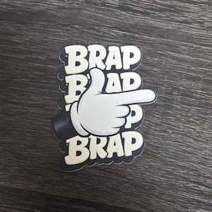 May include: A die-cut sticker featuring a white cartoon-style gloved hand pointing to the right. The background consists of the word BRAP repeated in a bold, cream-colored sans-serif font with a thick black outline, creating a layered effect.