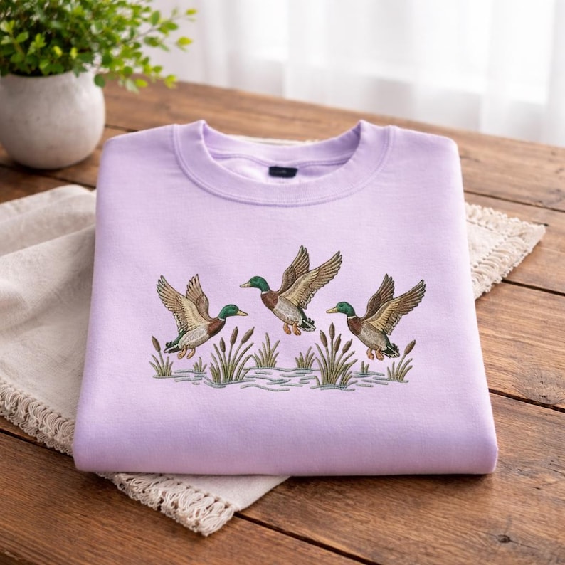 May include: A light purple sweatshirt featuring an embroidered design of three ducks in flight above reeds and water. The ducks have brown wings, green heads, and yellow beaks. The sweatshirt is made of a soft material and is suitable for casual wear.