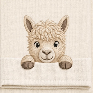 Op de afbeelding: Een crèmekleurige handdoek met een geborduurd ontwerp van een pluizige lamakop met donkerbruine oren en hoeven. De lama is zo geplaatst dat het lijkt alsof hij over de gevouwen rand van de handdoek kijkt, met gedetailleerd stikwerk.