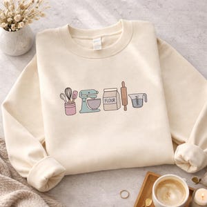 May include: Cream-coloured sweatshirt with a line of baking-themed illustrations. The design includes a mixer, flour bag, rolling pin, measuring jug, and a container with whisks and spatulas. The word "FLOUR" is printed on the flour bag.