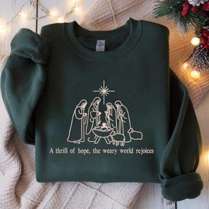 May include: Dark green sweatshirt with a white graphic of the Nativity scene, including the star, figures, and a baby in a manger. The text "A thrill of hope, the weary world rejoices" is printed below the graphic. The sweatshirt is folded.