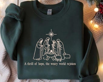 May include: Dark green sweatshirt with a white graphic of the Nativity scene, including the star, figures, and a baby in a manger. The text "A thrill of hope, the weary world rejoices" is printed below the graphic. The sweatshirt is folded.