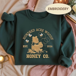 May include: Dark green sweatshirt with embroidered design. The design features a bear holding a honey pot, surrounded by bees, and the text "Hundred Acre Woods Honey Co. Est. 1926". The sweatshirt has long sleeves and a crew neck.