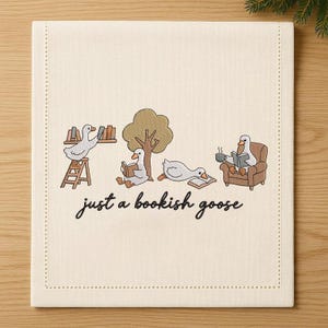 May include: Cream-colored linen table runner with an embroidered design featuring geese reading books. One goose is on a ladder, another under a tree, a third is lounging, and the last is in a chair. The text "just a bookish goose" is below.