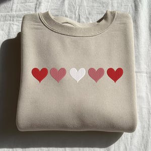 May include: A beige sweatshirt with a crew neck, featuring five embroidered hearts in a row. The hearts alternate in color: red, light pink, white, light pink, and red. The sweatshirt is folded neatly.