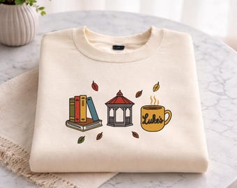 May include: Cream-colored sweatshirt with an embroidered design featuring books, a gazebo, and a coffee mug with the text "Luke's". The design includes autumn leaves. The sweatshirt is folded on a white surface.