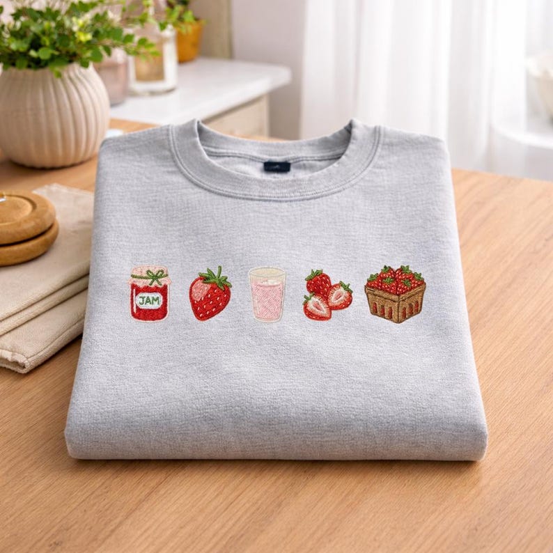 Puede incluir: Sudadera gris claro con dise&ntilde;os bordados de fresas. Los dise&ntilde;os incluyen un tarro de mermelada, una fresa entera, una bebida rosa en un vaso, fresas en rodajas y una cesta de fresas. La sudadera est&aacute; hecha de un material suave.