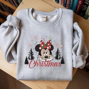 Op de afbeelding: Een lichtgrijs sweatshirt met een geborduurd Minnie Mouse-ontwerp, een rode strik, zuurstokken, kerstbomen en het woord "Christmas" in rode letters.