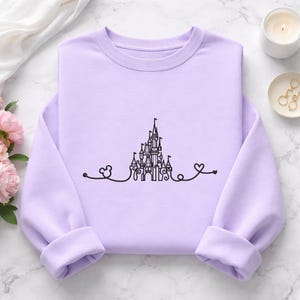 Puede incluir: Una sudadera lila con un dibujo lineal negro de un castillo, una cabeza de Mickey Mouse y dos corazones. La sudadera tiene cuello redondo y mangas largas con puños enrollados. El diseño está centrado en la parte delantera.