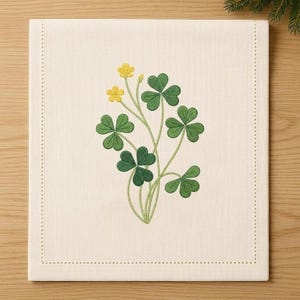 Könnte beinhalten: Cremefarbener quadratischer Tischset mit einem gestickten Design aus grünen Kleeblättern und gelben Blumen. Das Tischset hat einen genähten Rand. Die Kleeblätter und Blumen sind in einem naturalistischen Muster angeordnet.