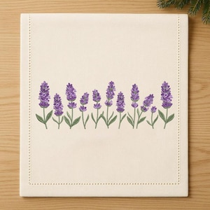 Puede incluir: Camino de mesa rectangular de lino color crema con una hilera de ramitas de lavanda bordadas. Las flores de lavanda son moradas, con tallos y hojas verdes. Los bordes del camino de mesa están acabados con puntada punteada.