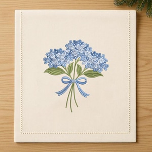 Può includere: Tovaglietta color crema con un design ricamato dettagliato di fiori di ortensia blu legati con un nastro blu. La tovaglietta ha un bordo cucito ed è posta su una superficie di legno.
