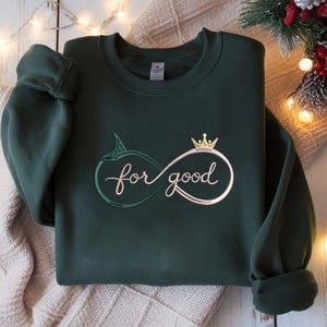 May include: Dark green sweatshirt with the phrase "for good" in a cursive font, integrated into an infinity symbol design. The infinity symbol is adorned with a crown. The sweatshirt is folded on a neutral-toned surface.
