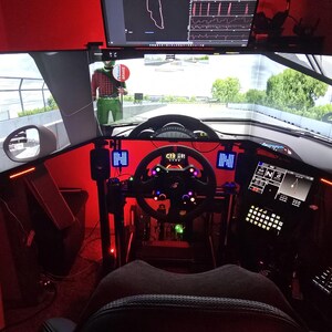 May include: A racing simulator setup featuring three large monitors displaying a track view, a central steering wheel with illuminated buttons, a digital dashboard display, and a mechanical keyboard, all bathed in a vibrant red ambient light.