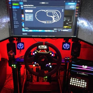 May include: A racing simulator setup featuring a black steering wheel with integrated buttons and a digital display, positioned in front of a triple monitor array showing a track map for a test drive, all illuminated by red ambient lighting.