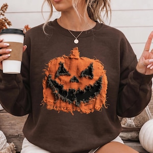 May include: A dark brown long-sleeve crewneck sweatshirt featuring a large, distressed orange pumpkin graphic with a jagged, dark-colored face. The pumpkin design has a textured, frayed fabric appearance, centered on the chest of the garment.