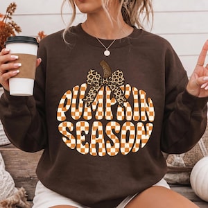 May include: A dark brown long-sleeve sweatshirt featuring a large graphic of a pumpkin with an orange and white checkered pattern. The pumpkin is topped with a leopard print bow and the text "PUMPKIN SEASON" is written across the center.