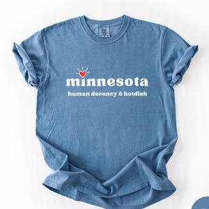May include: A blue jean colored t-shirt with the word "minnesota" in white, a red heart, and the phrase "human decency & hotdish" in white. The shirt is folded and has a tag at the neckline.