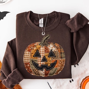 May include: A dark brown long-sleeve sweatshirt featuring a large, central pumpkin graphic made of various patchwork fabric patterns in shades of orange, brown, and tan, with black stitched eyes and a mouth, and a silver sunburst pendant necklace.