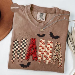 May include: An espresso-colored short-sleeve t-shirt featuring the word MAMA in large, patterned letters. Each letter has a unique design: black and white checkerboard, red leopard print, cherry print, and tan leopard print. Black bats and spiderwebs are printed around the text.