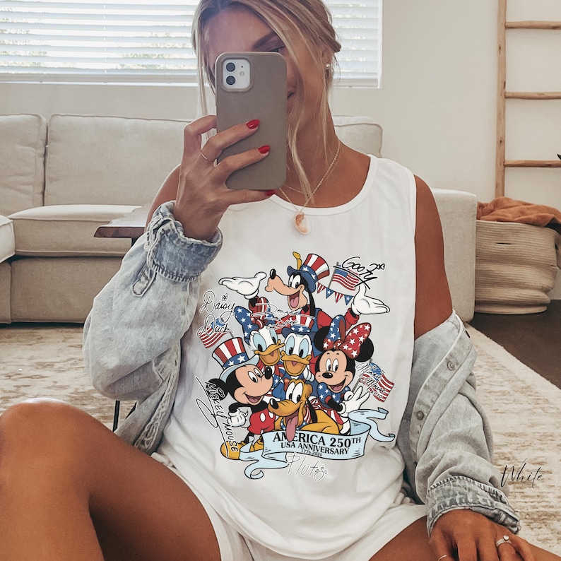 May include: A white sleeveless tank top featuring a colorful graphic print of Disney characters including Mickey Mouse, Minnie Mouse, Donald Duck, Daisy Duck, Goofy, and Pluto wearing patriotic hats, with text reading America 250th USA Anniversary.