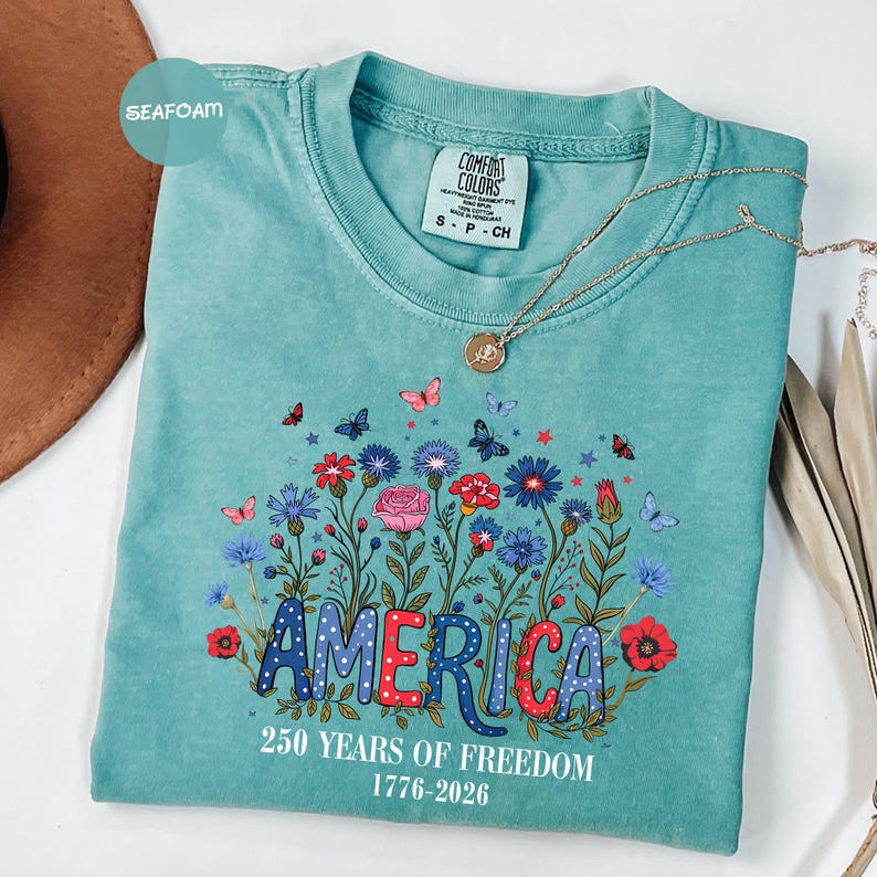 May include: Seafoam green t-shirt with a floral design and the word "AMERICA" in red, white, and blue. The shirt also features the text "250 YEARS OF FREEDOM 1776-2026". A gold necklace is draped across the shirt.