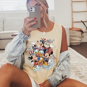 May include: A light yellow sleeveless tank top featuring a graphic print of various cartoon characters wearing patriotic red, white, and blue hats. The text reads America 250th USA Anniversary 1776-2026, with individual character names written in script.