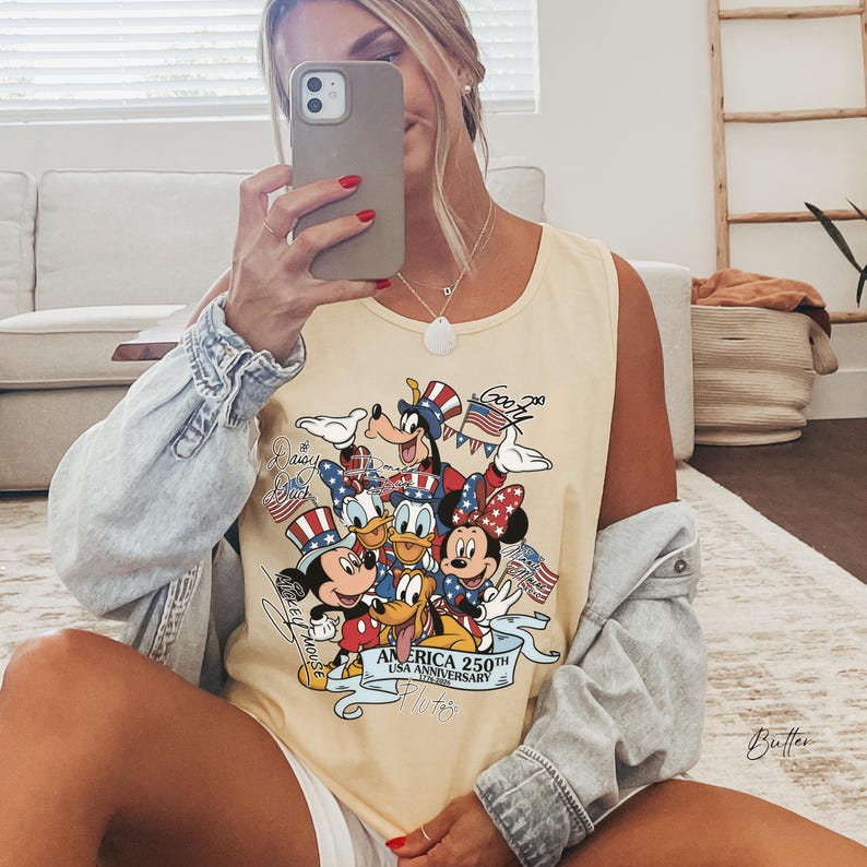 May include: A light yellow sleeveless tank top featuring a graphic print of various cartoon characters wearing patriotic red, white, and blue hats. The text reads America 250th USA Anniversary 1776-2026, with individual character names written in script.