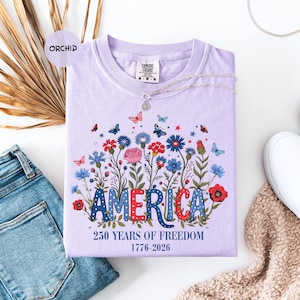 May include: A lilac-colored t-shirt with a floral design and the word "AMERICA" in red, white, and blue. The shirt also features the text "250 YEARS OF FREEDOM 1776-2026". A silver necklace with a pendant is draped across the shirt.