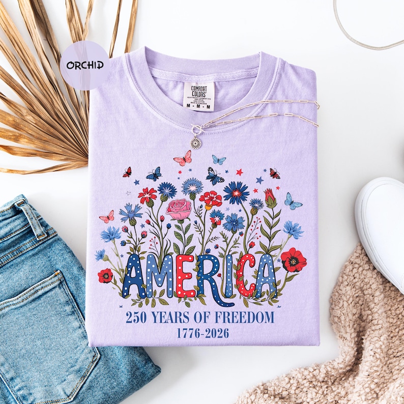 May include: A lilac-colored t-shirt with a floral design and the word "AMERICA" in red, white, and blue. The shirt also features the text "250 YEARS OF FREEDOM 1776-2026". A silver necklace with a pendant is draped across the shirt.