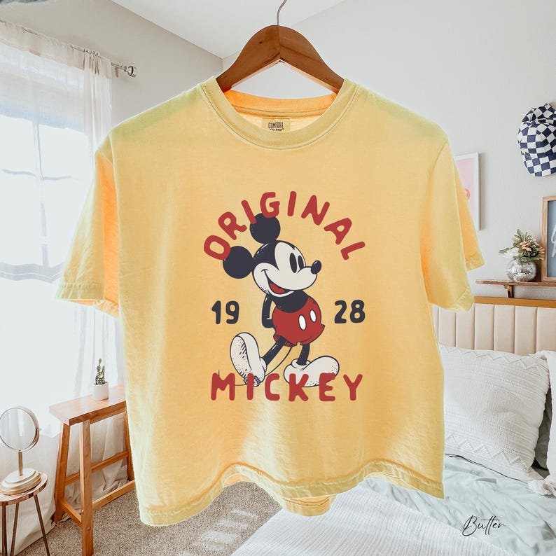 Puede incluir: Una camiseta de manga corta de color amarillo p&aacute;lido con un gr&aacute;fico de Mickey Mouse en el centro. El texto ORIGINAL aparece sobre el personaje y MICKEY debajo, con los n&uacute;meros 19 y 28 a los lados. El dise&ntilde;o utiliza tintas de color rojo y negro.