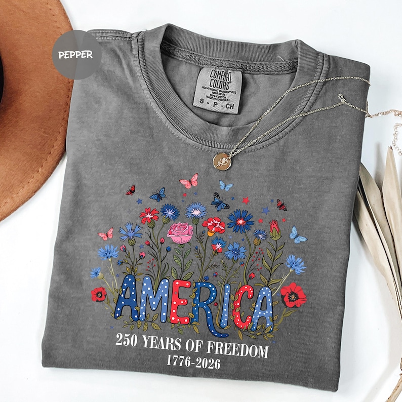 May include: A gray t-shirt with the word "AMERICA" in red and blue polka dot letters, surrounded by colorful flowers and butterflies. The text "250 YEARS OF FREEDOM 1776-2026" is below the word "AMERICA". A silver necklace is draped across the shirt.