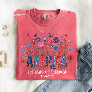 May include: A watermelon-colored t-shirt featuring the word "AMERICA" in red and blue, with a floral design and butterflies. The text "250 YEARS OF FREEDOM 1776-2026" is printed below. The shirt is folded on a white surface with jeans.