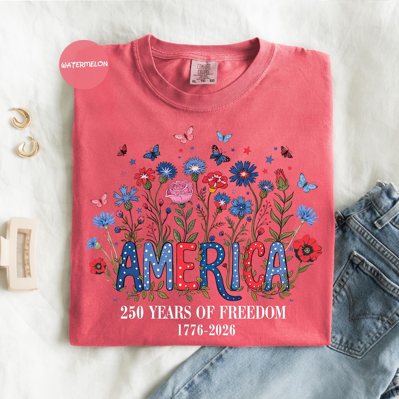 May include: A watermelon-colored t-shirt featuring the word "AMERICA" in red and blue, with a floral design and butterflies. The text "250 YEARS OF FREEDOM 1776-2026" is printed below. The shirt is folded on a white surface with jeans.