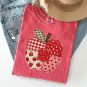 May include: A coral pink t-shirt featuring a large apple-shaped applique made of various fabric patches including red gingham, white with red apples, red with white flowers, and red hearts. The shirt has a Comfort Colors label at the neck.