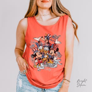 May include: A bright salmon sleeveless tank top featuring a graphic print of Disney characters including Mickey Mouse, Minnie Mouse, Donald Duck, Daisy Duck, Goofy, and Pluto wearing patriotic hats, with text reading America 250th USA Anniversary.