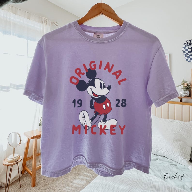 Puede incluir: Una camiseta de manga corta de color morado claro con un gr&aacute;fico de Mickey Mouse en el centro. El texto dice ORIGINAL MICKEY con los n&uacute;meros 19 y 28 a ambos lados del personaje. La camiseta tiene cuello redondo y un corte holgado.