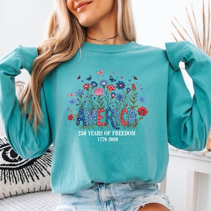 May include: Teal sweatshirt with the word "AMERICA" in red, white, and blue, surrounded by a floral design with butterflies. The text "250 YEARS OF FREEDOM 1776-2026" is below the word "AMERICA".