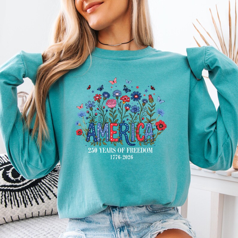 May include: Teal sweatshirt with the word "AMERICA" in red, white, and blue, surrounded by a floral design with butterflies. The text "250 YEARS OF FREEDOM 1776-2026" is below the word "AMERICA".