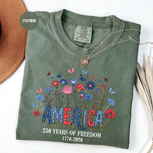May include: A moss green t-shirt with a floral design and the word "AMERICA" in red, white, and blue. The shirt also features the text "250 YEARS OF FREEDOM 1776-2026". A silver necklace with a pendant is draped across the shirt.