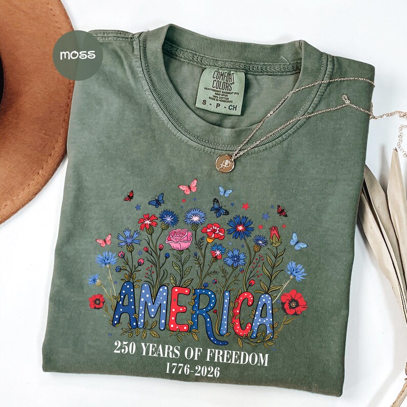 May include: A moss green t-shirt with a floral design and the word "AMERICA" in red, white, and blue. The shirt also features the text "250 YEARS OF FREEDOM 1776-2026". A silver necklace with a pendant is draped across the shirt.